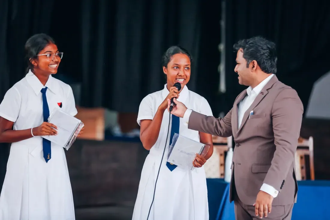 A student answers on the microphone, held for her by one of the speakers, while a classmate stands beside her with a workbook.
