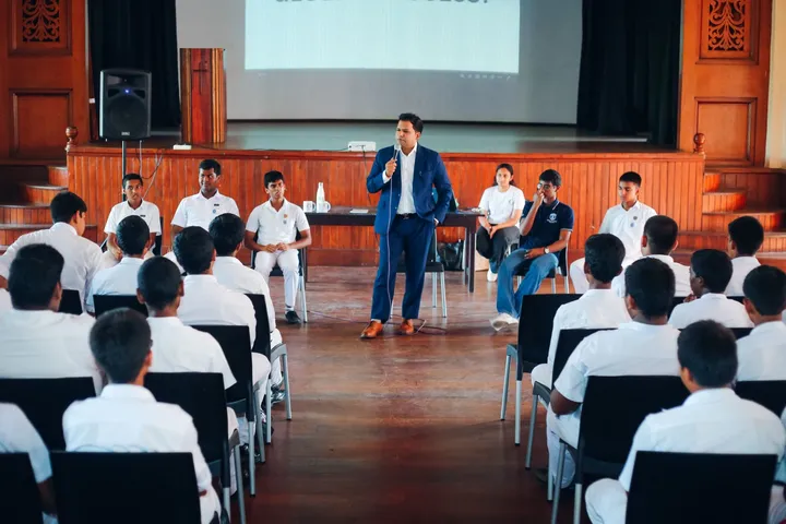 The school hall from the back, rows of students facing a speaker standing in the aisle.