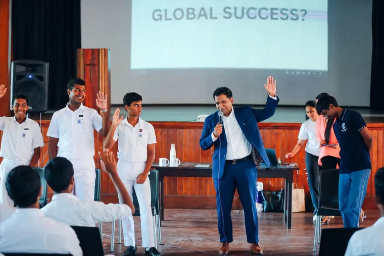 A speaker with a raised hand addresses the hall while several students stand with their hands up, a slide reading Global Success behind them.