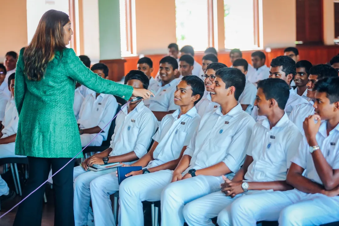 A speaker holds a microphone out towards a row of seated students, who are laughing and looking up at her.